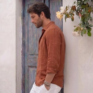 May include: A rust-colored linen shirt with long sleeves and a button-down collar. The shirt is paired with white pants. A multi-strand bracelet is visible on the wrist. The background includes a blue door and white wall with flowers.