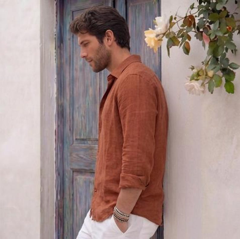 May include: A rust-colored linen shirt with long sleeves and a button-down collar. The shirt is paired with white pants. A multi-strand bracelet is visible on the wrist. The background includes a blue door and white wall with flowers.