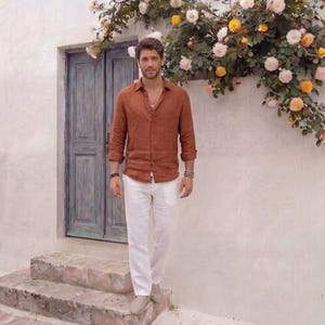 May include: A man wearing a rust-colored button-down shirt and white pants. He is standing on stone steps in front of a weathered blue door. The background features a white wall adorned with flowers.