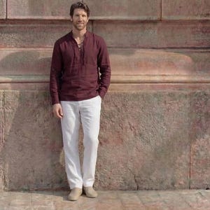 Puede incluir: Una camisa de manga larga en color granate oscuro con cuello acordonado y pantalones blancos. La camisa presenta un tejido texturizado y un dobladillo recto. Los pantalones son largos, de color blanco liso, con zapatos beige claros.