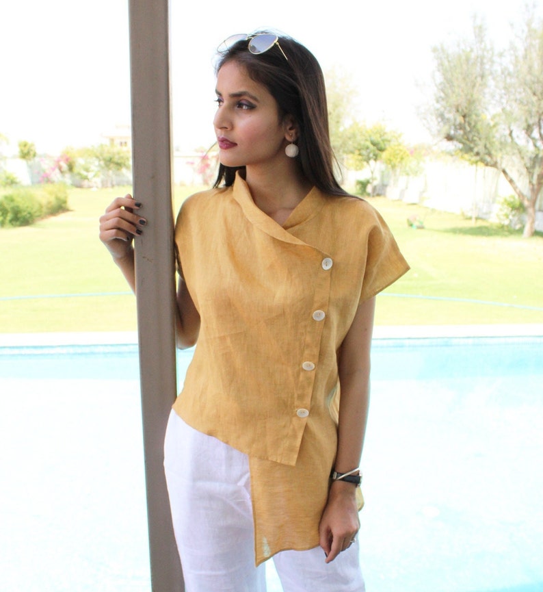 May include: A mustard yellow linen top with a diagonal button closure and a short, asymmetrical hem. The top has a collar and short sleeves.