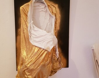 May include: A white fabric sculpture draped over a black canvas, with gold paint applied to the fabric. The sculpture is hanging on a wall.