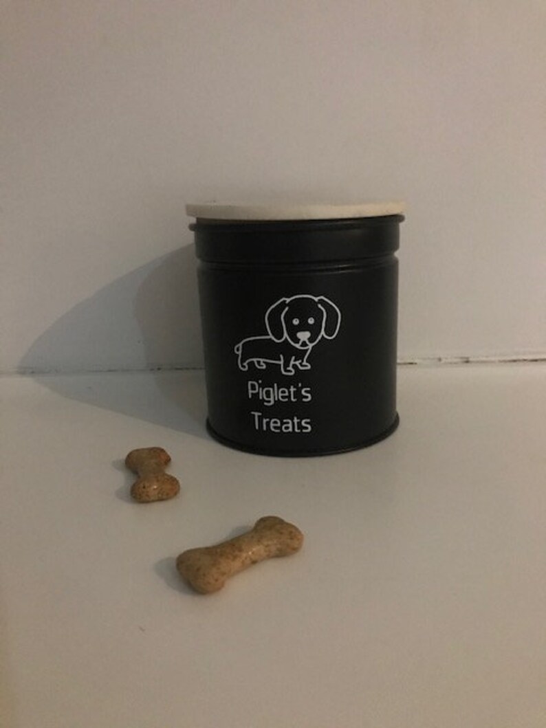 Pode incluir: Pote preto para guloseimas com tampa branca e uma silhueta branca de um dachshund. O pote diz "Piglet's Treats" na frente. Dois biscoitos para c&atilde;es est&atilde;o em primeiro plano.
