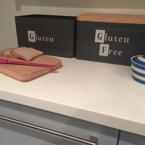 May include: Two grey bread bins with wooden lids. The bin on the left says "Gluten" and the bin on the right says "Gluten Free". There is a wooden chopping board with two slices of bread on it and a pink knife. There is also a blue and white striped butter dish.