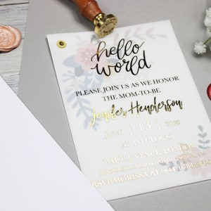 Op de afbeelding: Een witte babyshower-uitnodiging met gouden letters. De uitnodiging luidt "hello world" en bevat de tekst "Please join us as we honor the mom-to-be Jennifer Henderson, June 13, 2020, at 3 o'clock, Whole Venue, London. RSVP Morissa at +44 07812..."