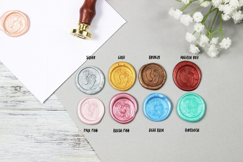 Op de afbeelding: Assortiment van waszegels in verschillende kleuren, waaronder zilver, goud, brons, antiek rood, lichtroze, blush roze, lichtblauw en turquoise. Elke zegel heeft een ontwerp met twee voetafdrukken.