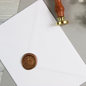 Op de afbeelding: Een witte envelop met een gouden waszegel met een voetafdrukdesign. Een rode waszegel met een voetafdrukdesign is ook zichtbaar op de achtergrond.