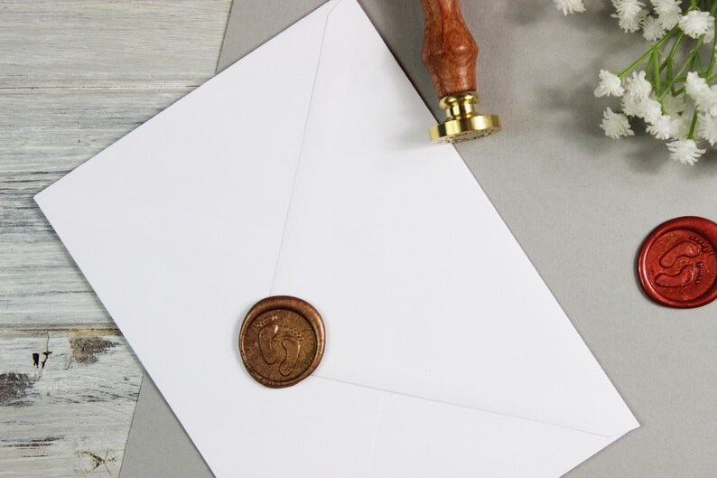 Op de afbeelding: Een witte envelop met een gouden waszegel met een voetafdrukdesign. Een rode waszegel met een voetafdrukdesign is ook zichtbaar op de achtergrond.