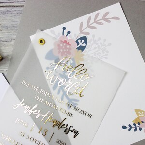 Op de afbeelding: Een doorschijnende witte uitnodiging met gouden folielettering die "hello world" en "Wordt u alstublieft bij ons om de aanstaande moeder Jennifer Thompson te eren op 13 juni 2020 om 15:00 uur!" luidt. De uitnodiging is versierd met bloemillustraties in roze, blauw en geel.