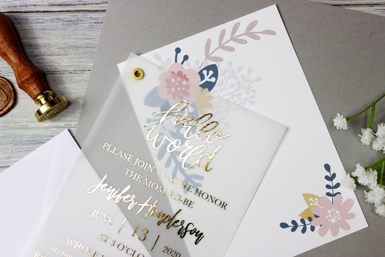 Op de afbeelding: Een doorschijnende witte uitnodiging met gouden folielettering die "hello world" en "Wordt u alstublieft bij ons om de aanstaande moeder Jennifer Thompson te eren op 13 juni 2020 om 15:00 uur!" luidt. De uitnodiging is versierd met bloemillustraties in roze, blauw en geel.