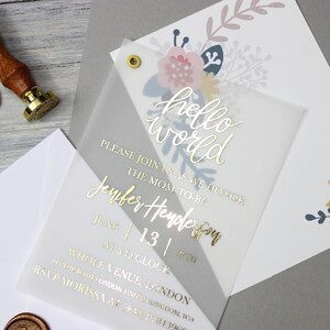 Op de afbeelding: Een transparante babyshower-uitnodiging met gouden letters die "hello world" luidt. De uitnodiging bevat ook de tekst "Sluit je bij ons aan om de aanstaande moeder, Jennifer Henderson, te eren op 13 juni om 15:00 uur. Whole Venue, Londen. RSVP Morissa op +44 1284 3960". De uitnodiging is versierd met een bloemmotief in roze, blauw en goud.