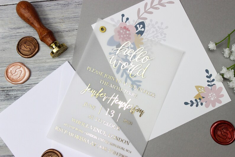 Op de afbeelding: Een transparante babyshower-uitnodiging met gouden letters die "hello world" luidt. De uitnodiging bevat ook de tekst "Sluit je bij ons aan om de aanstaande moeder, Jennifer Henderson, te eren op 13 juni om 15:00 uur. Whole Venue, Londen. RSVP Morissa op +44 1284 3960". De uitnodiging is versierd met een bloemmotief in roze, blauw en goud.