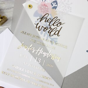 Op de afbeelding: Een witte en grijze uitnodiging met gouden folielettering die "hello world" en "Wordt u alstublieft bij ons om de aanstaande moeder Jennifer Henderson te eren op 13 juni 2020 om 15:00 uur op de hele locatie, locatie" zegt.
