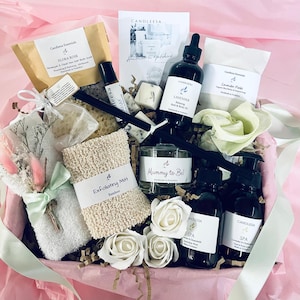May include: A gift basket filled with bath and body products, including a bath mitt, soap, body oil, and a small bottle of essential oil. The basket is decorated with white roses and a small bouquet of flowers. The basket is labelled "Mummy to Be!" and includes the text "CandlEesa Handmade in Ephemeris".