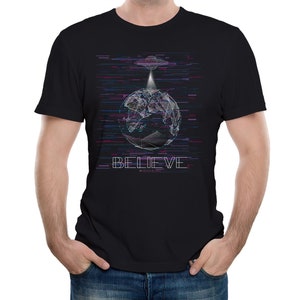 Puede incluir: Una camiseta negra con un gráfico de un globo terráqueo con un OVNI encima. El gráfico está en un estilo pixelado con un efecto de glitch. El texto "BELIEVE" está debajo del gráfico.