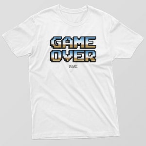 Op de afbeelding: Wit t-shirt met een gepixeld "GAME OVER" ontwerp in blauw, goud en bruin. Het shirt heeft ook de tekst "REALITY GJ33CH-I" eronder. Een casual t-shirt voor dagelijks gebruik.
