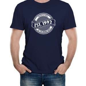Op de afbeelding: Een marineblauw t-shirt met een witte cirkelvormige afbeelding met de tekst "Perfectly Aged Est. 1992 All The Original Parts".