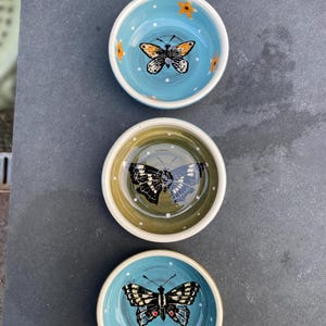 May include: Three small, round ceramic bowls with butterfly designs. The bowls are painted in shades of blue and olive green, with white polka dots and butterfly illustrations. The butterflies have detailed wing patterns and are in black, orange, and white.