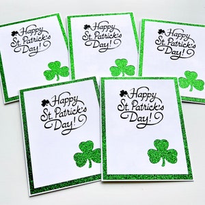 May include: Five blank greeting cards with a green glitter border and shamrock design. The cards read "Happy St. Patrick's Day!" in black cursive lettering.