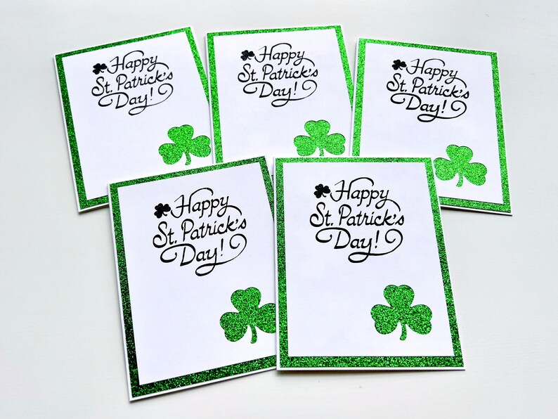 May include: Five blank greeting cards with a green glitter border and shamrock design. The cards read "Happy St. Patrick's Day!" in black cursive lettering.
