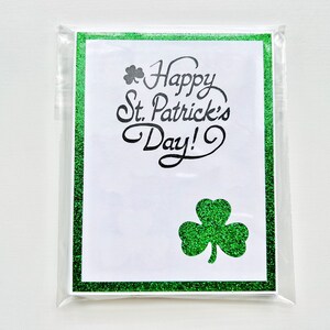 May include: A white card with a green glitter border and shamrock design. The card reads "Happy St. Patrick's Day!" in black cursive lettering.