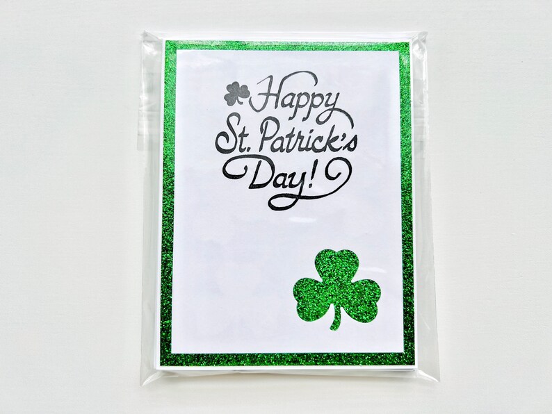 May include: A white card with a green glitter border and shamrock design. The card reads "Happy St. Patrick's Day!" in black cursive lettering.