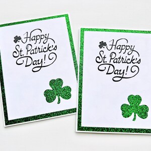 May include: Two white greeting cards with green glitter borders. Each card has a shamrock and the text "Happy St. Patrick's Day!" in black.
