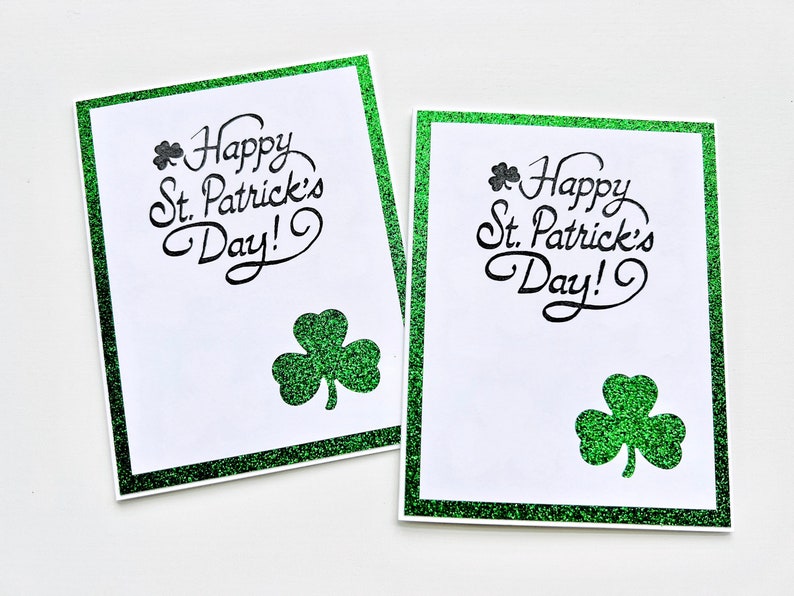 May include: Two white greeting cards with green glitter borders. Each card has a shamrock and the text "Happy St. Patrick's Day!" in black.
