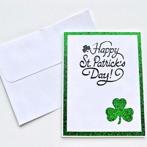 May include: A green glitter card with a white border and the text "Happy St. Patrick's Day!" in black script. A small shamrock is above the text and a larger shamrock is below the text.