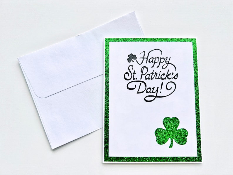 May include: A green glitter card with a white border and the text "Happy St. Patrick's Day!" in black script. A small shamrock is above the text and a larger shamrock is below the text.