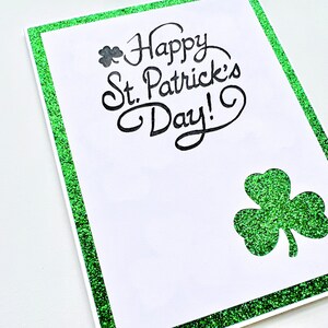 May include: A white card with a green glitter border. The card says "Happy St. Patrick's Day!" in black cursive writing. A green glitter shamrock is in the bottom right corner.