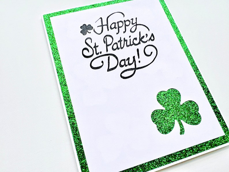 May include: A white card with a green glitter border. The card says "Happy St. Patrick's Day!" in black cursive writing. A green glitter shamrock is in the bottom right corner.