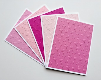 May include: A set of six blank greeting cards with a heart pattern in shades of pink and magenta. The cards are arranged in a fan shape.