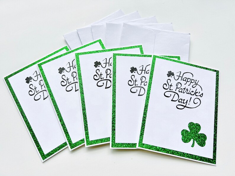 May include: Six blank greeting cards with a green glitter border and the text "Happy St. Patrick's Day!" on one card. The cards are white with a shamrock design on the card with the text.