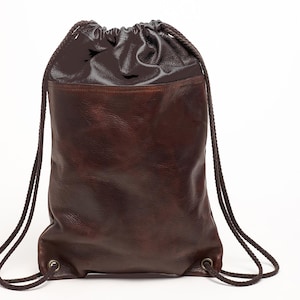 Puede incluir: Mochila con cordón de cuero marrón oscuro. La parte superior es brillante, la inferior mate. Tiene cordones de cuerda marrón oscuro y un diseño clásico. La mochila está vacía y sobre un fondo blanco.