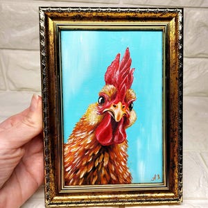 Puede incluir: Pintura enmarcada de un gallo con cresta y barbilla rojas, sobre un fondo azul claro. Las plumas del gallo son de tonos naranja y marr&oacute;n. El marco es dorado con detalles ornamentados. Las iniciales del artista est&aacute;n en la esquina inferior derecha.