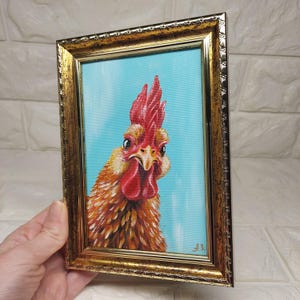 Puede incluir: Pintura enmarcada de un gallo con cresta y barbilla rojas, sobre un fondo azul claro. Las plumas del gallo son de tonos marrones y amarillos, y el marco es dorado con detalles ornamentados. La obra est&aacute; firmada en la esquina inferior derecha.