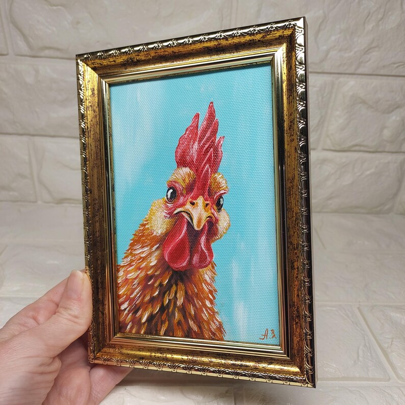 Puede incluir: Pintura enmarcada de un gallo con cresta y barbilla rojas, sobre un fondo azul claro. Las plumas del gallo son de tonos marrones y amarillos, y el marco es dorado con detalles ornamentados. La obra est&aacute; firmada en la esquina inferior derecha.