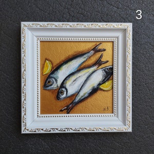 May include: A framed painting featuring three silver fish arranged diagonally on a metallic gold background, accompanied by two yellow lemon wedges. The white frame has gold decorative accents and the artist's initials A.B. are in the corner.