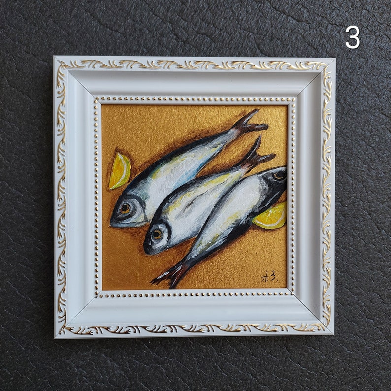 May include: A framed painting featuring three silver fish arranged diagonally on a metallic gold background, accompanied by two yellow lemon wedges. The white frame has gold decorative accents and the artist's initials A.B. are in the corner.