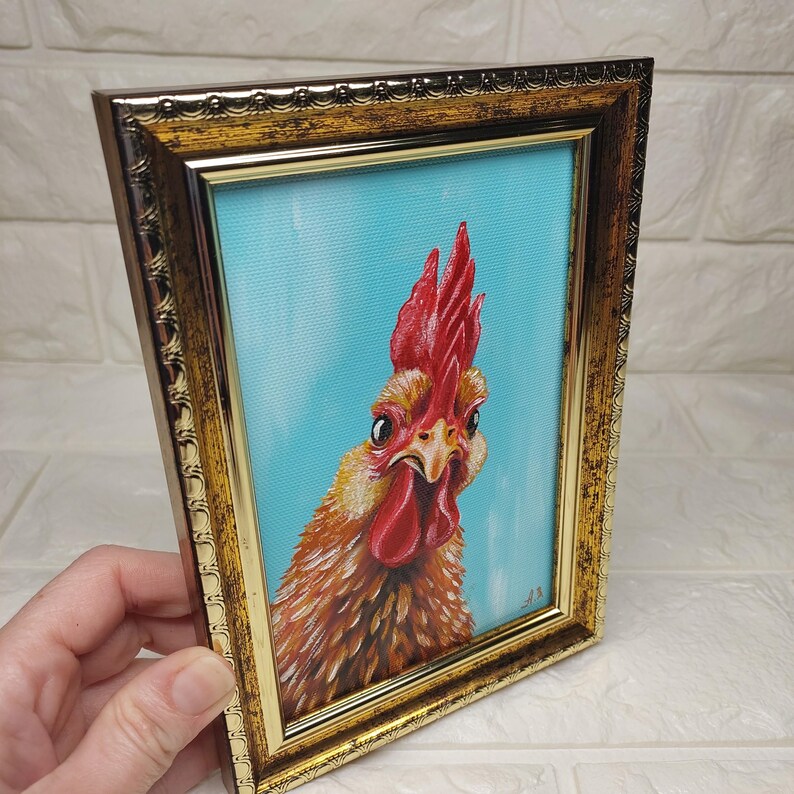 Puede incluir: Pintura enmarcada de un gallo con cresta y barbilla rojas, sobre un fondo azul claro. La obra de arte est&aacute; en un marco dorado ornamentado, creando una est&eacute;tica vintage. La cara del gallo es el punto focal.