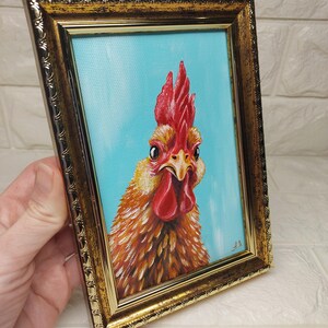 Puede incluir: Pintura enmarcada de un gallo con cresta y barbilla rojas, sobre un fondo azul claro. Las plumas del gallo est&aacute;n representadas en tonos naranja y marr&oacute;n, y el marco es dorado.