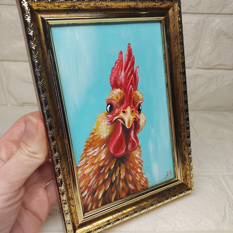 Puede incluir: Pintura enmarcada de un gallo con cresta y barbilla rojas, sobre un fondo azul claro. Las plumas del gallo est&aacute;n representadas en tonos naranja y marr&oacute;n, y el marco es dorado.