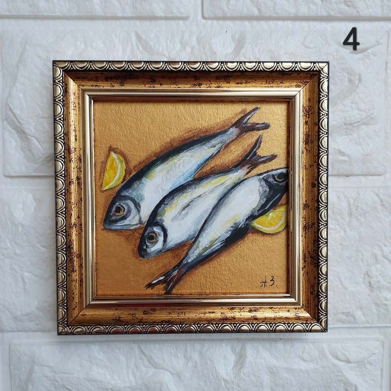 May include: A square framed painting featuring three silver fish arranged diagonally on a textured gold background, accompanied by two yellow lemon wedges. The frame is ornate, metallic gold with a dark, distressed finish and a scalloped edge.