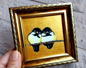 May include: A small square painting in a decorative gold frame featuring two birds with black heads and yellow and white bodies perched together on a thin brown branch against a solid metallic gold background.