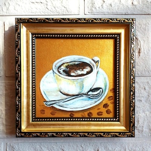 May include: A framed painting of a white coffee cup and saucer on a gold background. The cup is filled with dark coffee and a silver spoon rests on the saucer. Scattered coffee beans are painted on the surface. The frame is ornate gold and black.