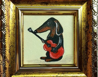 May include: A small square painting of a black and brown dachshund playing a bright red acoustic guitar. The artwork is set against a cream-colored background and is enclosed in an ornate, metallic gold frame with a textured, scalloped edge.