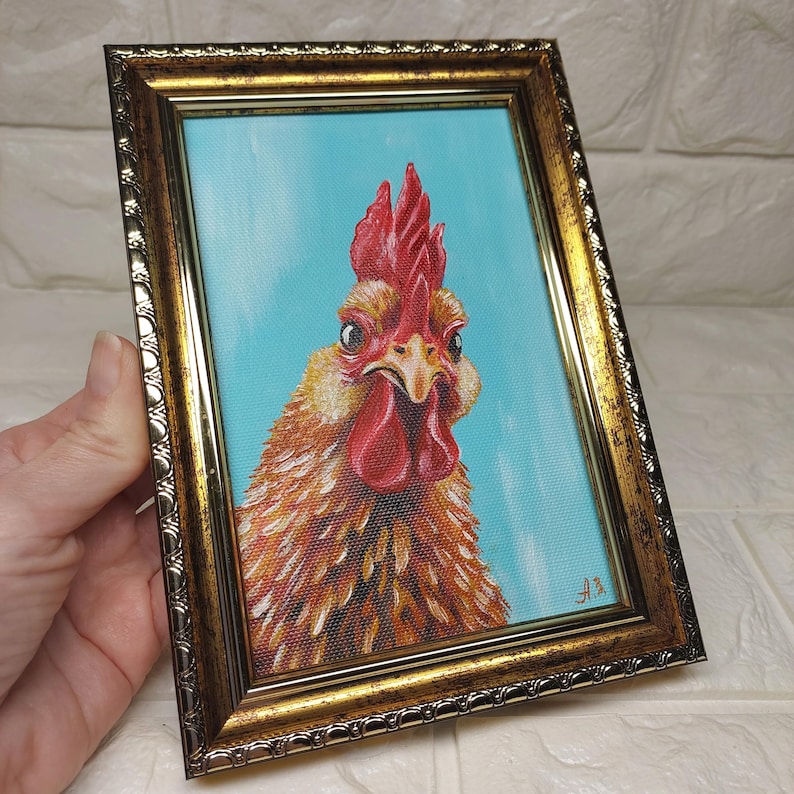 Puede incluir: Pintura enmarcada de un gallo con cresta y barbilla rojas, sobre un fondo azul claro. Las plumas del gallo son de tonos marrones y dorados. El marco es ornamentado con un acabado dorado.