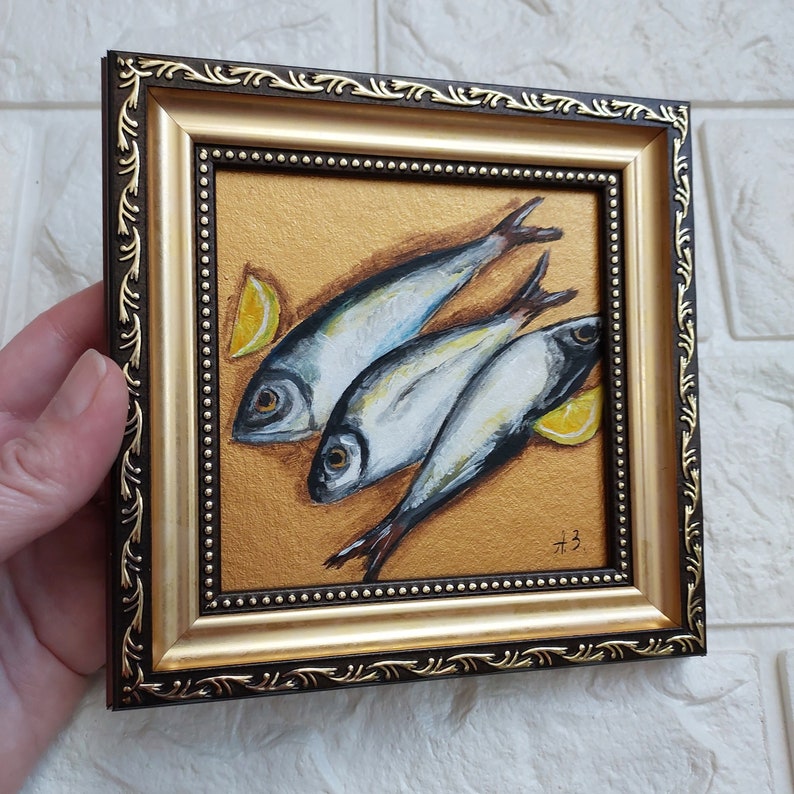 May include: A square framed painting featuring three silver fish arranged diagonally on a textured gold background, accompanied by two yellow lemon wedges. The frame is dark brown with ornate gold filigree detailing and a beaded inner border.