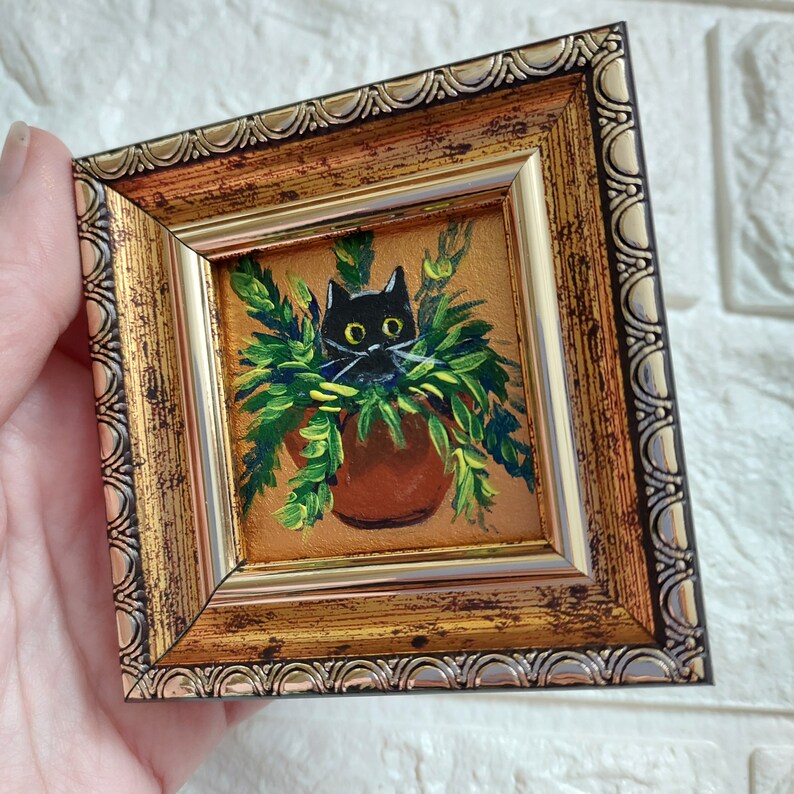 May include: A black cat with yellow eyes peeking out of a pot of green plants. The painting is framed in a gold ornate frame.